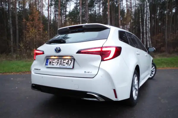 Biała Toyota Corolla 1.8 Hybrid na tle jesiennego lasu. Widok z tyłu, z podświetlonymi światłami.