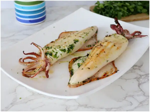 Due calamari alla griglia, conditi con erbe aromatiche, serviti su un piatto bianco.