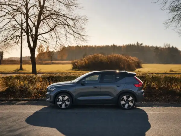 Szary Volvo EX40 zaparkowany na poboczu drogi, w tle jesienny krajobraz z drzewami i krzewami.