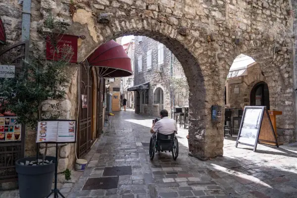 Wąska, brukowana uliczka w **Budva stare miasto**. Mężczyzna na wózku inwalidzkim przejeżdża pod kamiennym łukiem. Po bokach sklepy i restauracje.
