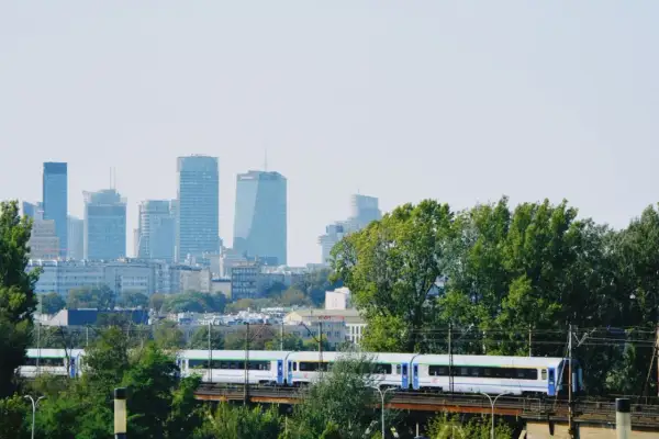 Pociąg PKP jedzie przez zielone drzewa z widokiem na warszawskie wieżowce. Szukasz tanich biletów PKP Warszawa - Kraków?