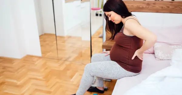 Kobieta w 27 tygodniu ciąży siedzi na łóżku, masując bolące plecy.