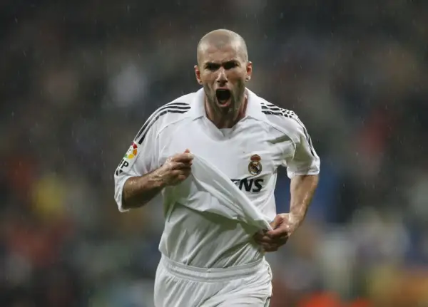Zinedine Zidane w białej koszulce Realu Madryt, z podniesioną koszulką, krzyczy radośnie do kibiców.