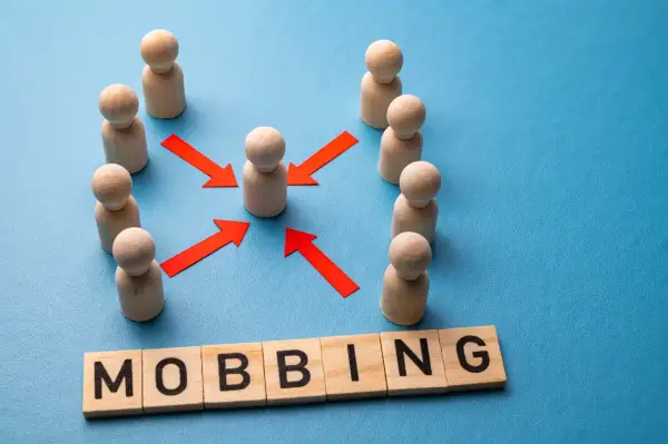 Drewniane figurki ludzi otaczają jedną, a czerwone strzałki wskazują na nią. Słowo "MOBBING" ułożone z liter. Jak udowodnić mobbing w pracy?