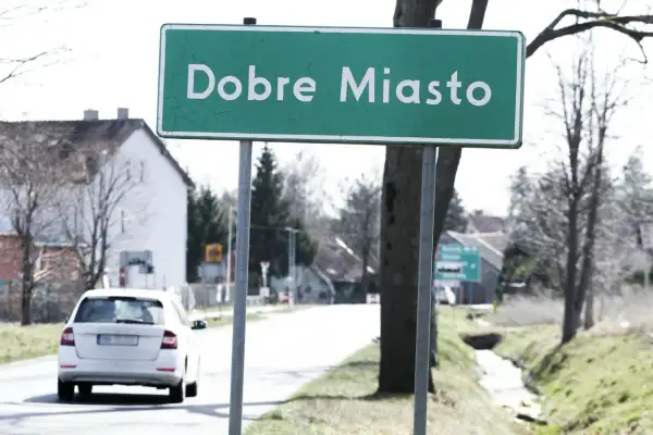 Zielony znak miejscowości "Dobre Miasto" wita podróżnych. W tle biały samochód i domy.