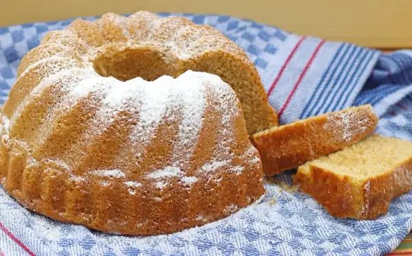 Puszysta babka z owocami, oprószona cukrem pudrem, czeka na spróbowanie. Obok leżą dwa kawałki ciasta.
