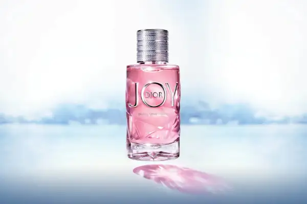 Różowy flakon Dior Joy 90ml z metalową nakrętką, na tle rozmytego, błękitnego krajobrazu.