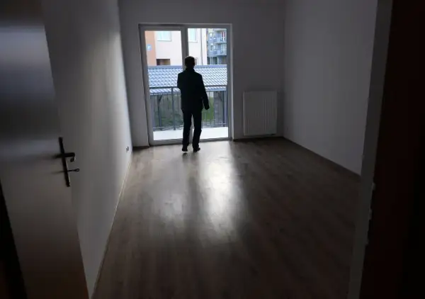 Mężczyzna w pustym mieszkaniu, zastanawia się, gdzie złożyć wniosek o mieszkanie komunalne. Widok na balkon i sąsiednie budynki.