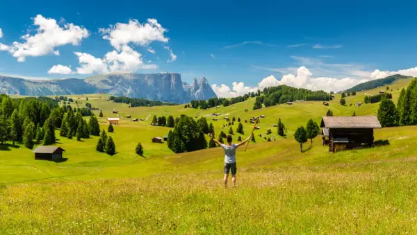 Ein Wanderer genießt die Aussicht auf eine Alm mit Berghütten und Wäldern. Perfekt für eine Mehrtageswanderung in den Dolomiten.