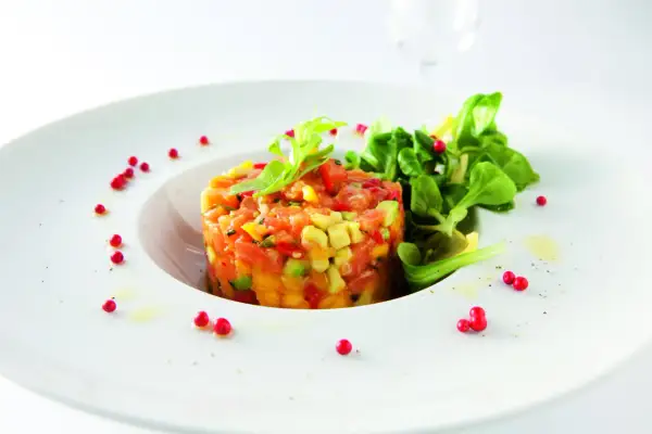 Délicieux tartare de truite aux agrumes, avocat et poivrons, garni de jeunes pousses et de baies roses sur une assiette blanche.