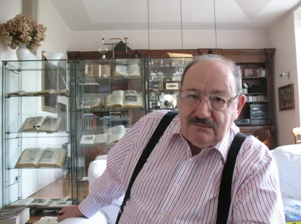 Wilhelm z Baskerville, w okularach i koszuli w paski, siedzi w otoczeniu antycznych ksiąg i gabinetów pełnych skarbów.