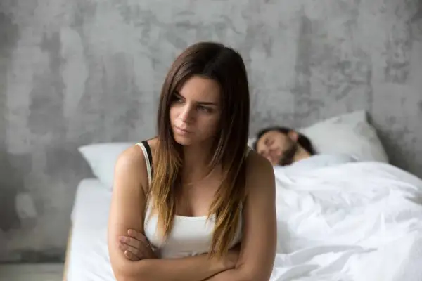Een vrouw zit met gekruiste armen, gefrustreerd, terwijl haar partner slaapt. Ze probeert zich in te leven in anderen, maar voelt zich alleen.