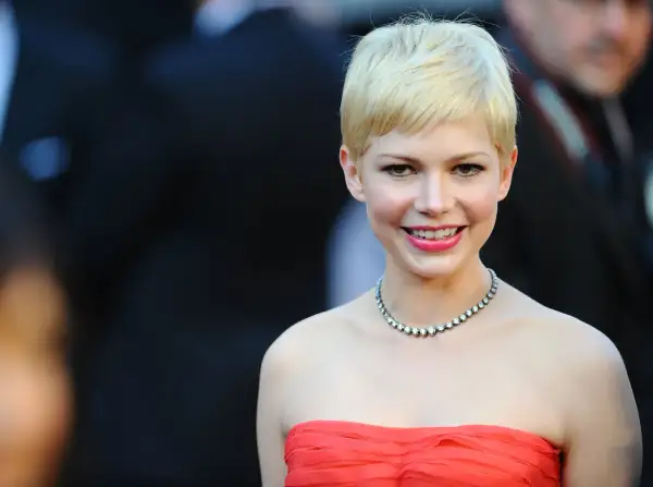 Eine Frau mit einem frechen Pixie-Cut und welligem Haar lächelt charmant. Sie trägt ein auffälliges Collier und ein rotes Kleid.