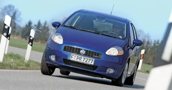 Niebieski Fiat Punto Grande pokonuje zakręt na drodze, z kierowcą w okularach za kierownicą.
