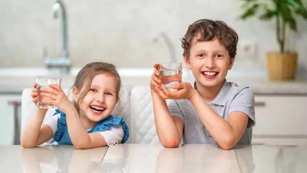 Uśmiechnięta dziewczynka i chłopiec trzymają szklanki z wodą. Cieszą się orzeźwiającym smakiem, jakby pili miękką wodę prosto ze źródła.