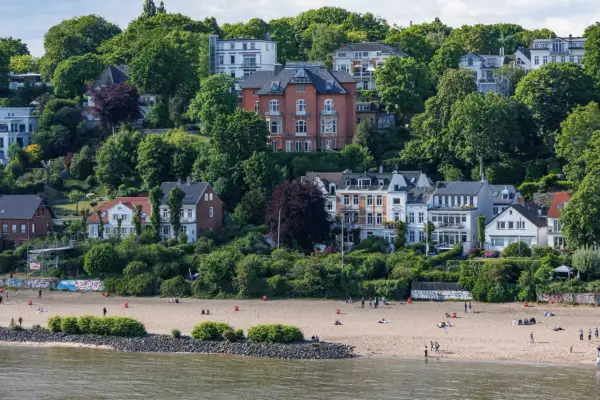 Prächtige Villen und grüne Hügel über einem Sandstrand an den Elbvororten Hamburgs. Menschen genießen den sonnigen Tag am Wasser.