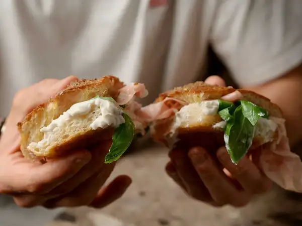En person håller en delad focaccia med krämig ost, skinka och färsk basilika. En härlig focaccia pesto-macka!