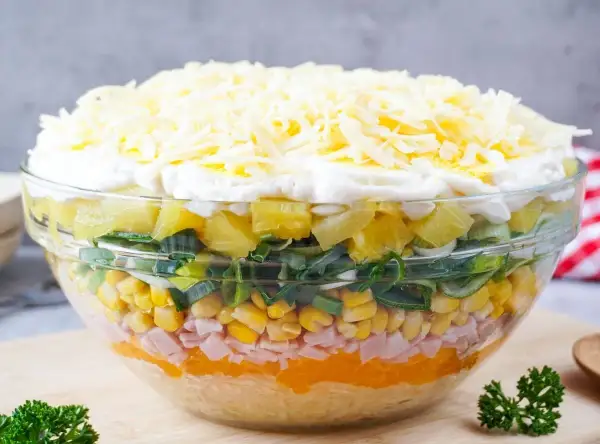 Schichtsalat mit Eisbergsalat, Mais, Schinken, Ananas, Lauch und Käse in einer Glasschüssel.