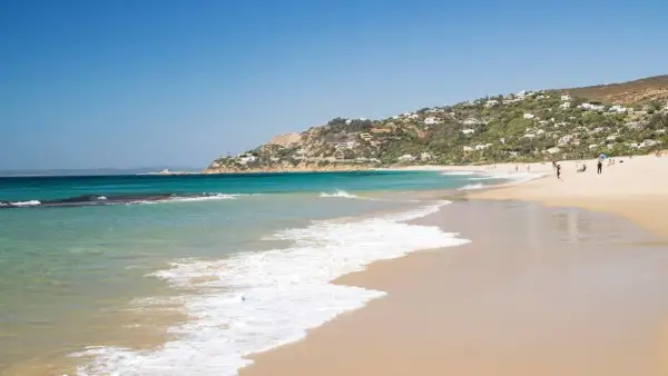 Aguas turquesas y arena dorada en las calas de Tarifa, con casas blancas salpicando la ladera verde. Un día perfecto para disfrutar del mar.