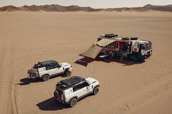 Un camion Dakar et deux 4x4 blancs dans le désert. L'équipe se réunit sous un auvent, prête pour l'aventure.