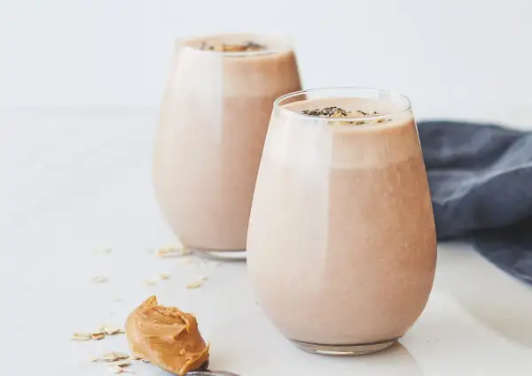 Två glas med krämig choklad- och jordnötssmoothie, toppad med chiafrön. En sked med jordnötssmör och havregryn ligger bredvid. En perfekt start på dagen, som Don Pedro skulle ha gillat.