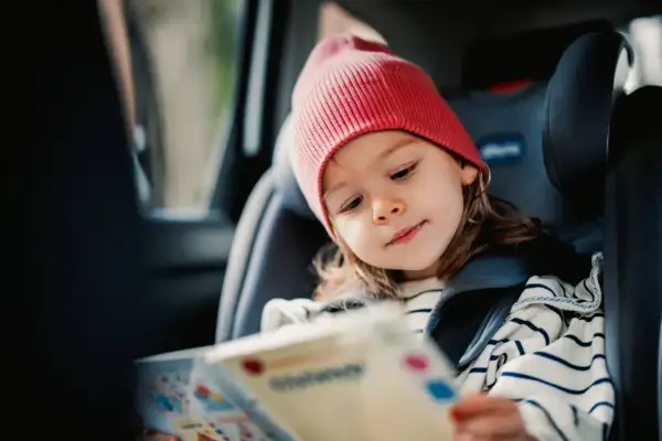 Kleines Mädchen mit roter Mütze liest ein Buch im Kindersitz. Perfekt für autofahrt spiele, um die Reise kurzweilig zu gestalten.
