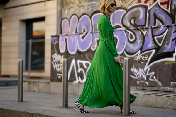 Donna con vestito lungo elegante verde smeraldo e scarpe zebrate, cammina con passo deciso davanti a un muro con graffiti.