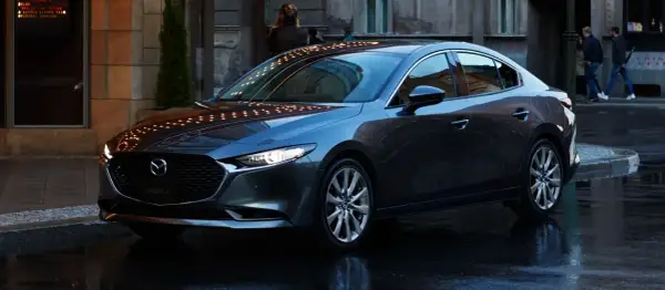 Elegancka, ciemnoszara Mazda 3 sedan stoi na mokrej ulicy, odbijając światła miasta.