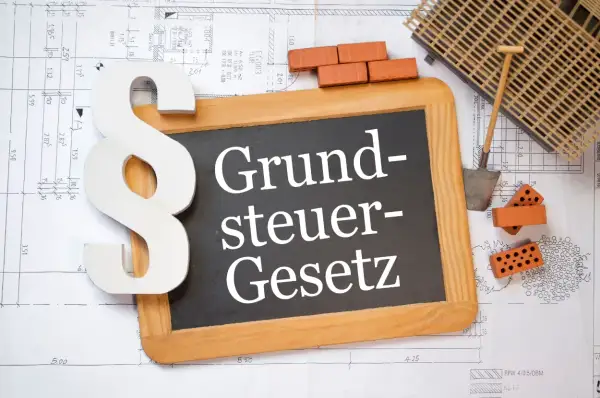 Ein Paragrafensymbol und eine Tafel mit "Grundsteuer-Gesetz" liegen auf Bauplänen. Ziegel und Werkzeug deuten auf den Grunderwerbsteuer in Niedersachsen hin.