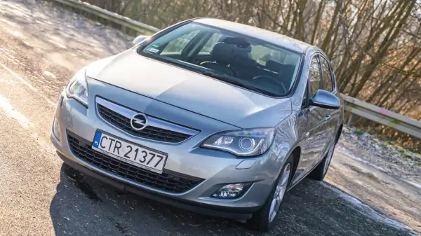 Srebrny Opel Astra J z przodu, zaparkowany na drodze z drzewami w tle.