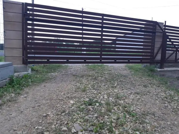 Brązowa, metalowa brama przesuwna z poziomymi szczeblinami. Widoczna jest szczelina pod bramą, przez którą widać trawę i żwir.