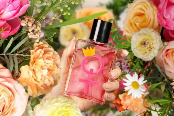 Dłoń trzyma flakonik z różowym płynem i symbolem Wenus zwieńczonym koroną. Czy perfumy z feromonami działają? Otoczenie bujnych kwiatów sugeruje zapach.
