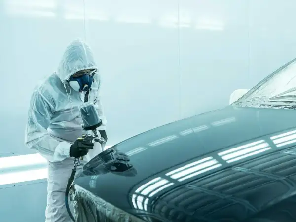 Pracownik w kombinezonie ochronnym wykonuje matowienie lakieru na samochodzie za pomocą pistoletu natryskowego.