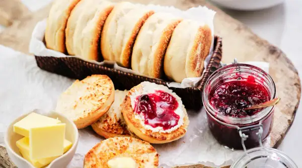 En korg med gyllene engelska muffins, en burk sylt och smör. En muffin är delad och bredd med smör och sylt.