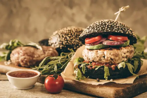 Gotowy hamburger z czarną bułką, kotletem wołowym i warzywami, obok surowy kotlet i sos.
