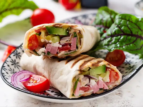Pyszna tortilla z szynką, awokado, pomidorami i serem feta, podana z sałatką i czerwoną cebulą.