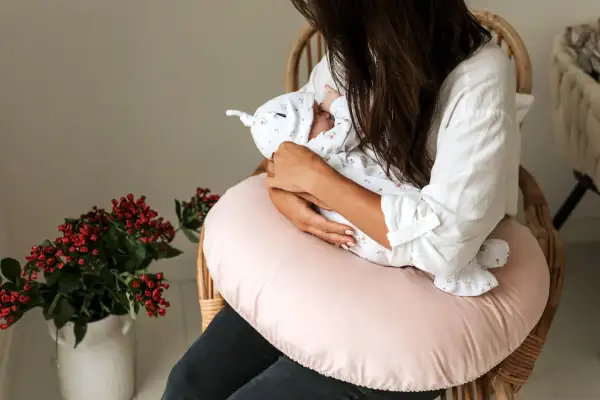Kobieta siedzi na krześle, trzymając na rękach noworodka. Pokazuje, jak karmić noworodka, otulając go delikatnie.