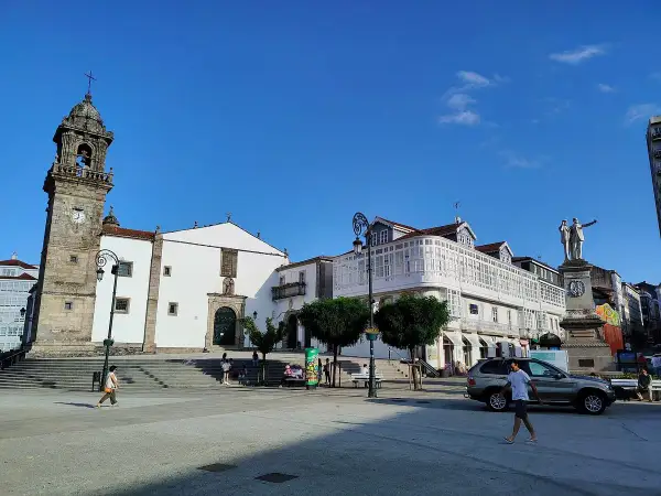 La plaza Betanzos, con su iglesia de torre campanario, edificios blancos y un monumento, bajo un cielo azul.