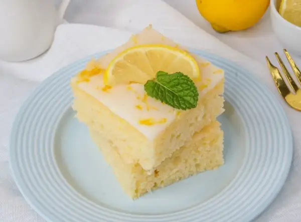 Zwei Stücke zitronenkuchen mit joghurt, garniert mit Zitronenscheibe und Minze, auf einem hellblauen Teller.