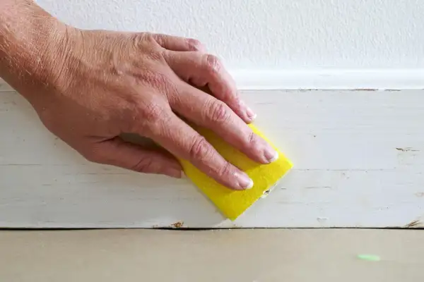 Hand med gul svamp slipar en vitmålad list. Förberedelser för att måla tunt.