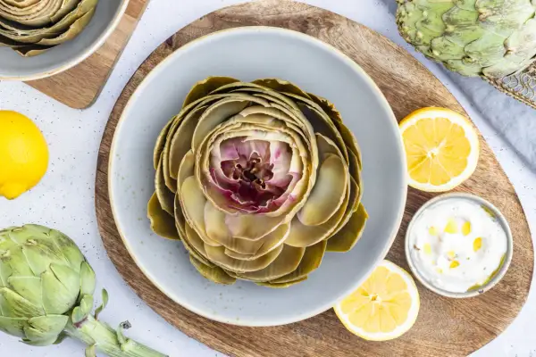Ein gekochter Artischocke auf einem Teller, bereit für ein leckeres Artischocken Rezept. Dazu Zitronenspalten und eine Dip-Schale.