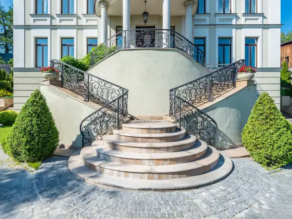 Imponente villa con scale esterne coperte da ringhiere in ferro battuto e ornate da vasi con fiori rossi.