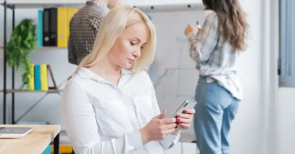 Kobieta z blond włosami korzysta z telefonu, być może używając wyszukiwania głosowego, podczas gdy koledzy pracują przy tablicy.
