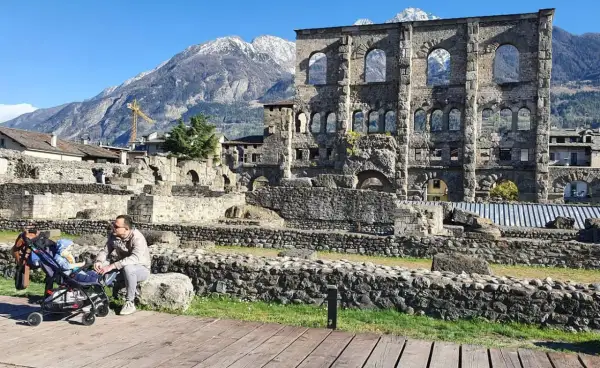 Un uomo con un passeggino ammira le rovine romane, uno dei **paesi più belli della Valle d'Aosta**, con montagne innevate sullo sfondo.