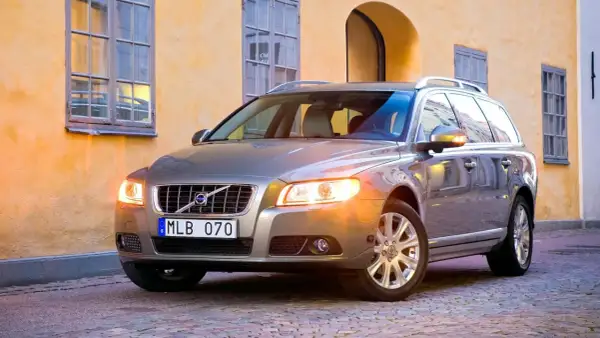 En servad bil, en silverfärgad Volvo kombi, står parkerad på en kullerstensgata framför en gul byggnad med fönster.