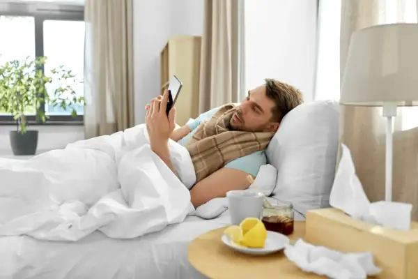 Chory mężczyzna w łóżku, owinięty kocem, przegląda telefon. Obok kubek z herbatą, cytryna i miód. To zwolnienie siła wyższa.