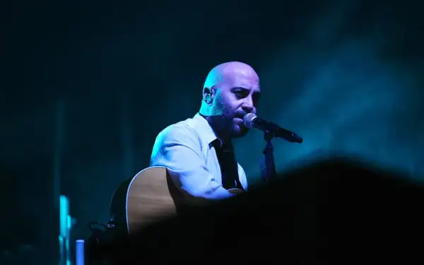 Giuliano Sangiorgi dei Negramaro, con la sua chitarra, canta al microfono, un passo indietro sul palco illuminato da luci blu.
