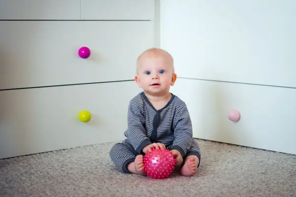 7 miesięczne dziecko z niebieskimi oczami siedzi na dywanie i bawi się różową, kolczastą piłeczką.