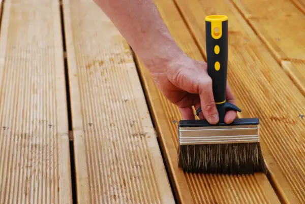 Main d'une personne utilisant une large brosse pour nettoyer bois après ponçage. La terrasse en bois est prête pour le traitement.