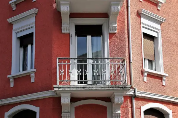 Dettaglio di un muro esterno con finitura a rasatura, finestre con persiane e un piccolo balcone con ringhiera in ferro battuto.
